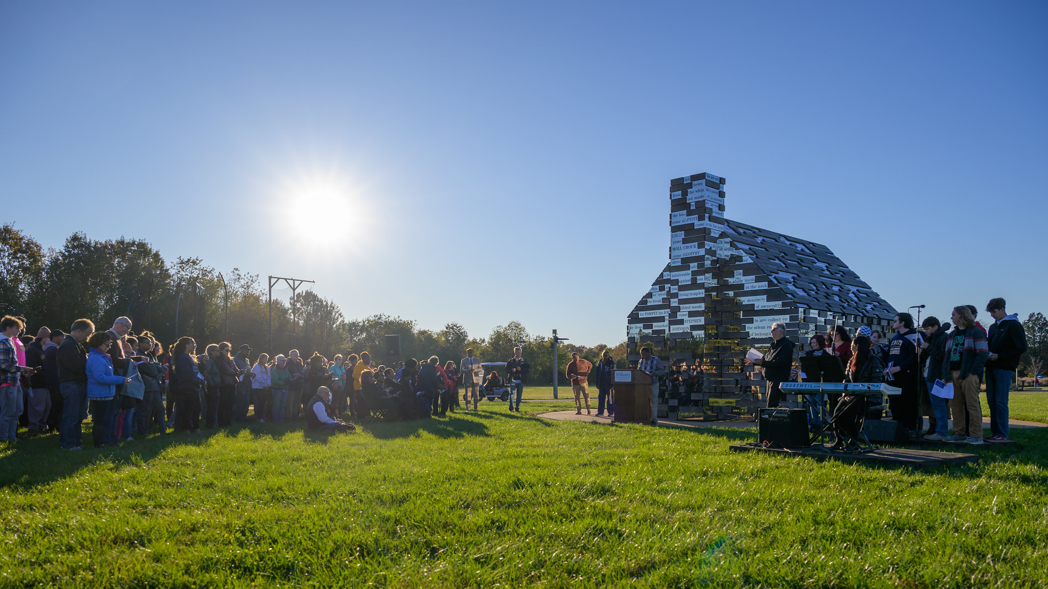 A crowd gathers on grass near a reflective, modern structure shaped like a house, with people performing music under a clear sky and bright sun.