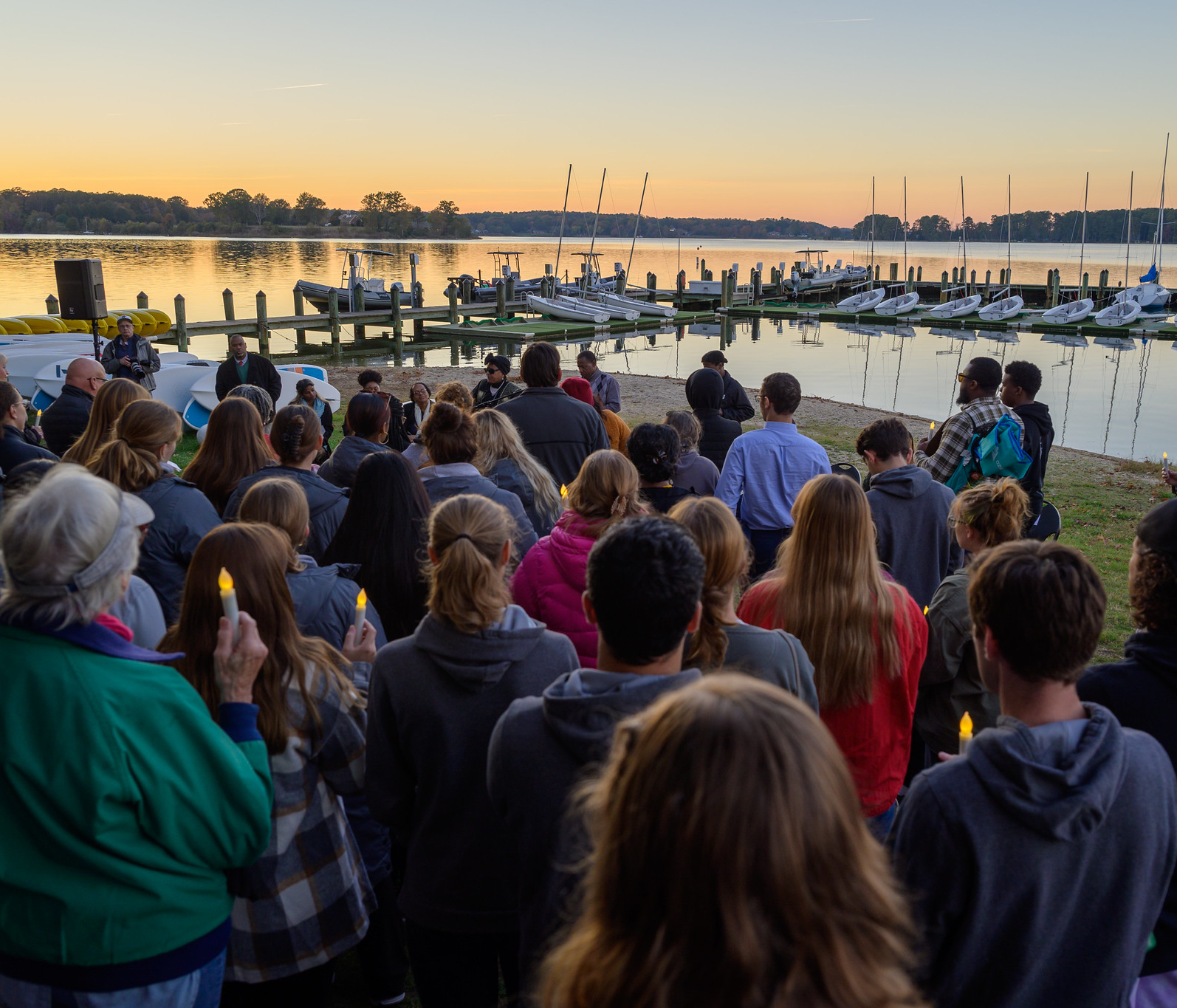 A group of people hold candles at a gathering by a marina at sunset, with sailboats and a calm lake in the background.