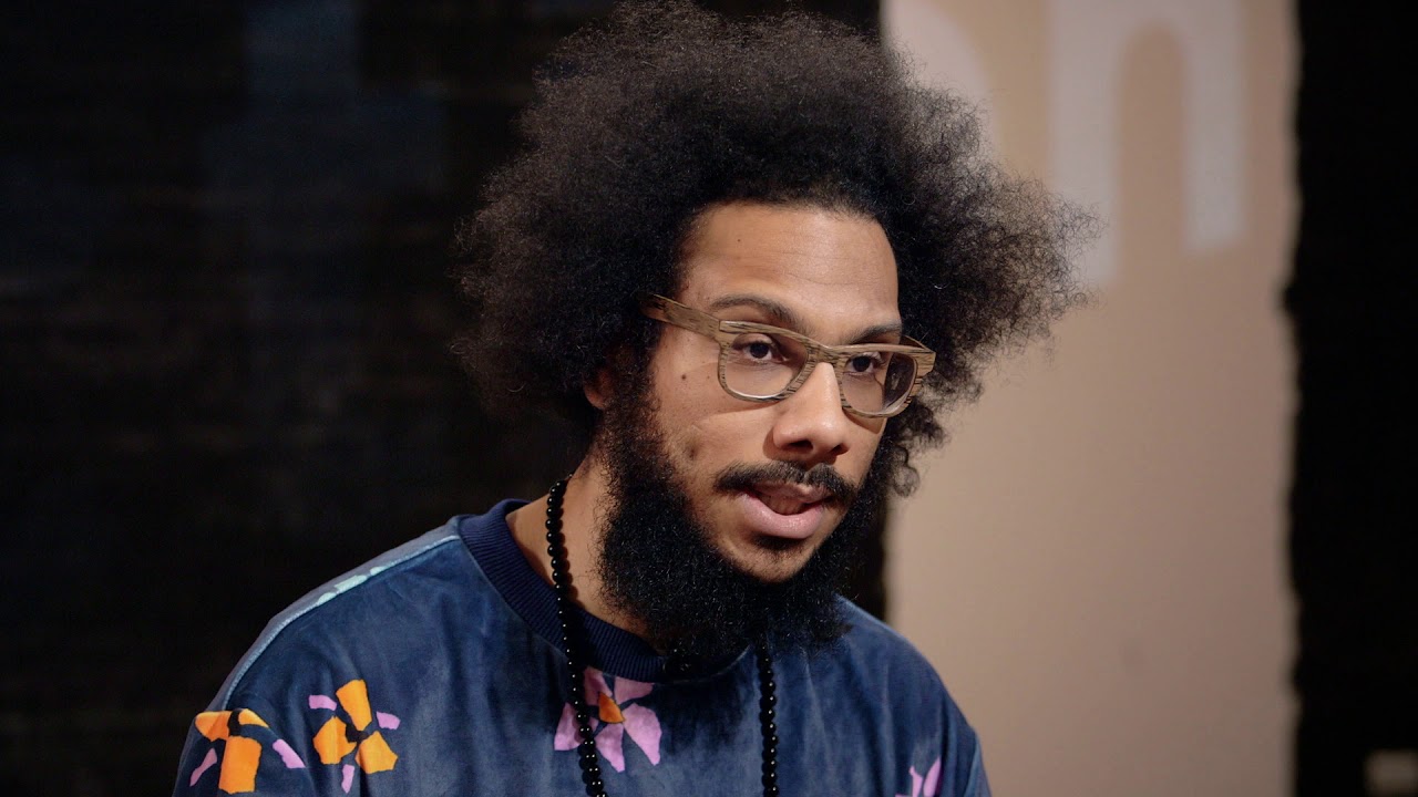 A man with glasses, an afro hairstyle, and a beard is wearing a blue floral-patterned shirt and a beaded necklace, facing slightly to the right.