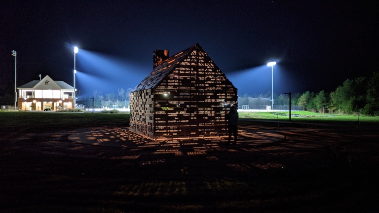 A small house-shaped sculpture with illuminated text cutouts stands on a field at night, with bright floodlights and a building visible in the background.