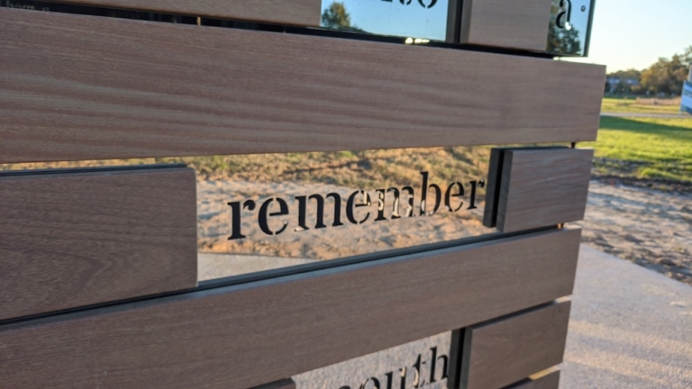 Wooden slats with the word "remember" cut out, sunlight casting shadows, and grass visible in the background.
