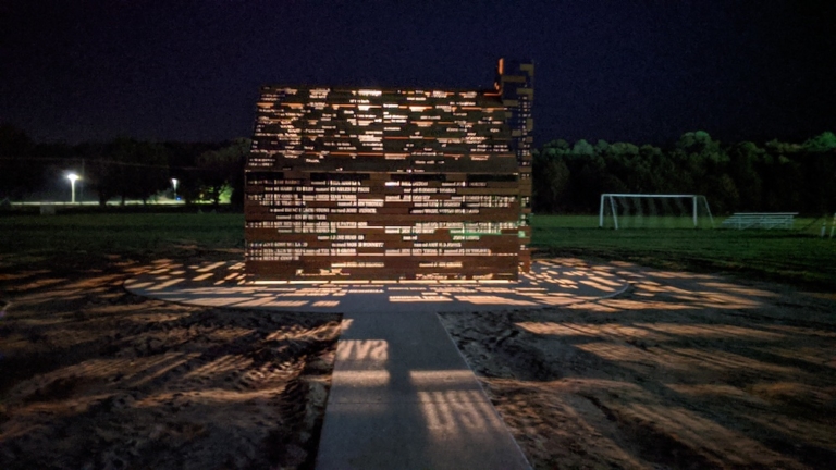 A small structure made of perforated metal panels is illuminated at night, casting patterned light and shadows onto the ground in a grassy field near a soccer goal.