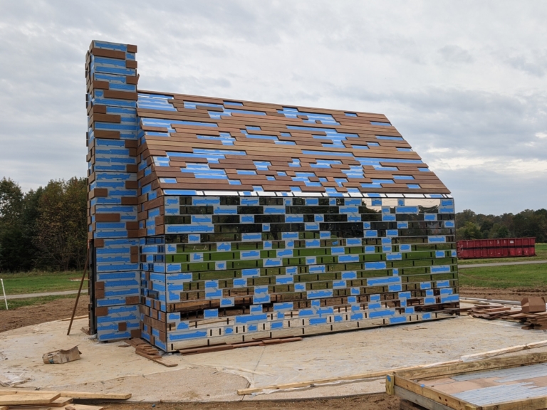 A small house structure with walls made of alternating blue, brown, and mirrored rectangular panels, standing on a concrete base in an open field.