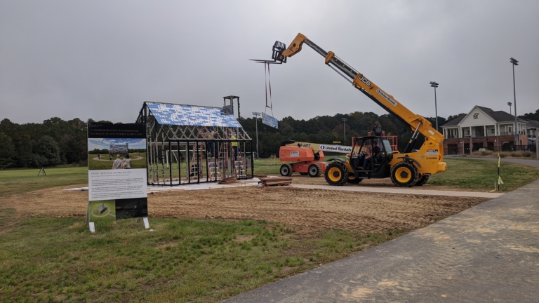 A telescopic forklift lifts a beam to workers assembling the frame of a small building on a construction site with nearby equipment and signage.