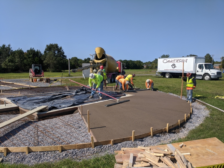Construction workers are leveling freshly poured concrete on a curved section of ground at an outdoor site, with trucks and equipment visible in the background.
