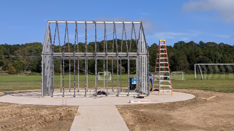 A metal frame structure under construction stands on a circular concrete base in a field, with a ladder and tools nearby. Soccer goals and trees are visible in the background.