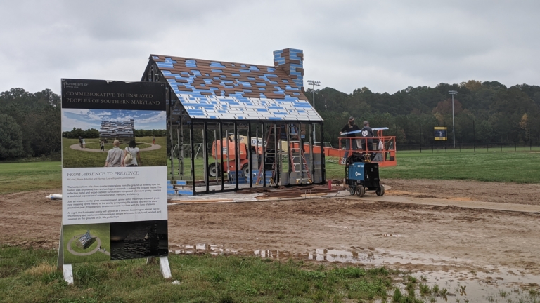 A partially constructed building stands in a field, with workers on a lift nearby and an informational sign about commemorating enslaved peoples in the foreground.
