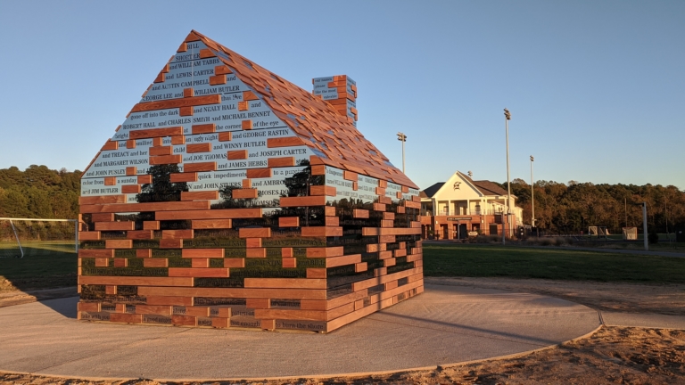 A small wooden house structure made from multicolored planks with engraved text, situated outdoors on a concrete pad with a sports field and building in the background.