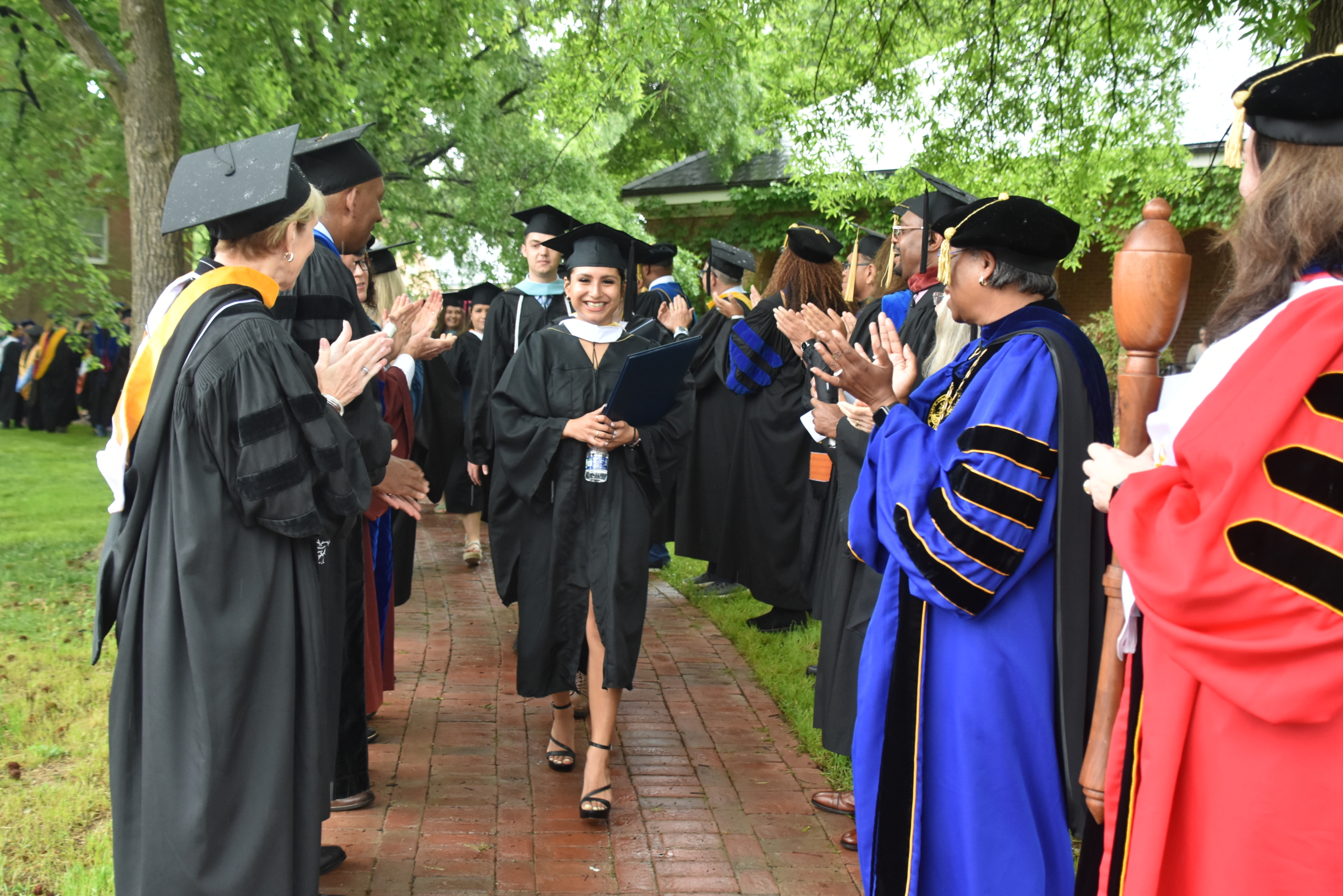 A graduate in a cap and gown walks happily down a brick pathway. Faculty members in academic regalia stand on either side, clapping and smiling. Trees line the background, and the atmosphere is celebratory.