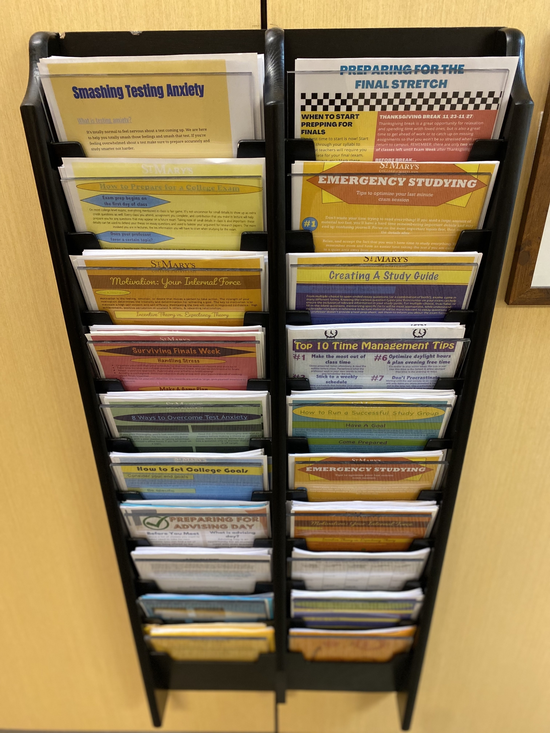 A wall-mounted rack holds a variety of colorful pamphlets, each with distinct titles related to study tips, exam preparation, time management, and college success strategies. The pamphlets are neatly organized in rows.
