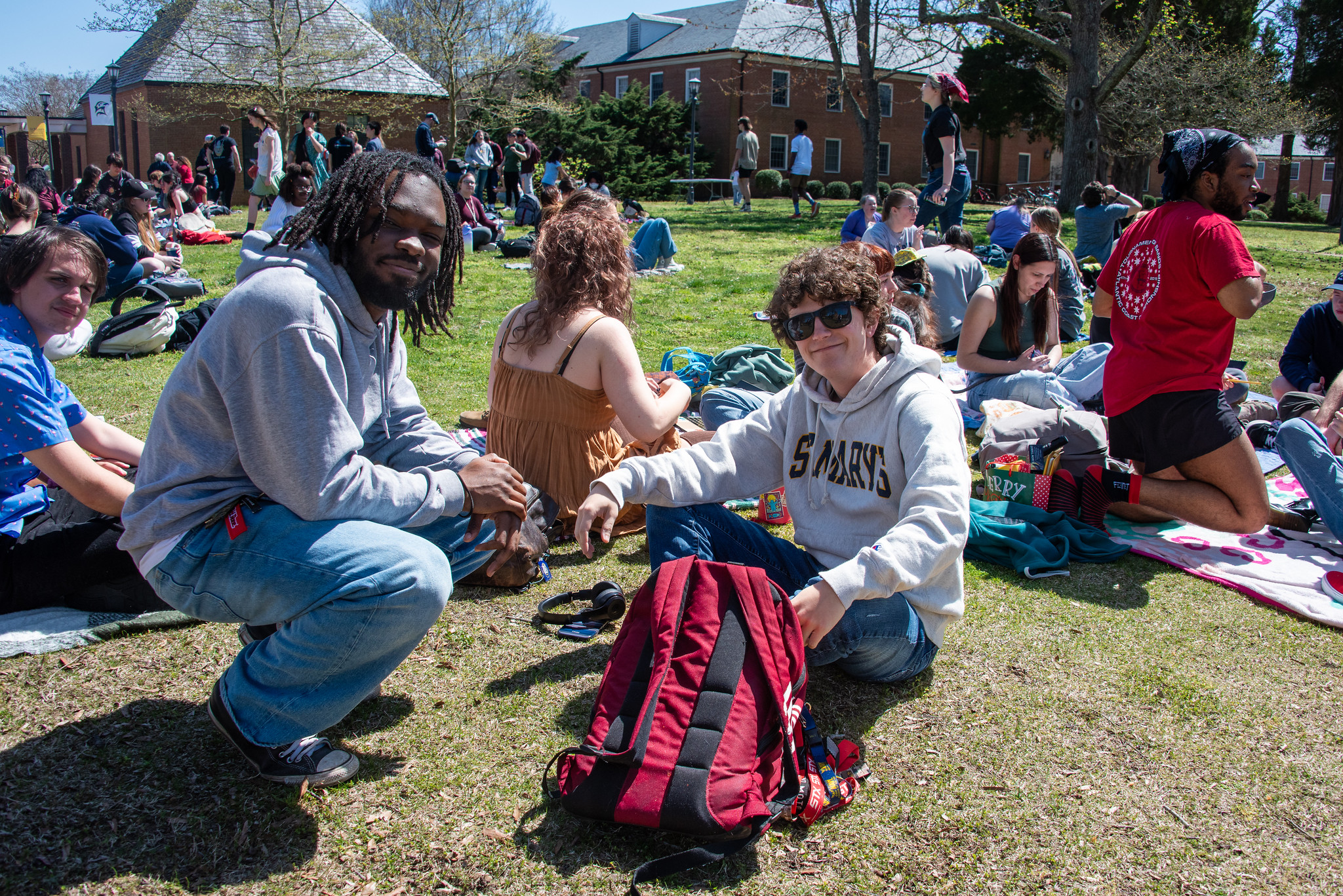 A group of people sitting on grass in a park-like setting on a sunny day. Two individuals sit in the foreground with a red and black backpack. Others are lounging or talking in the background. Trees and brick buildings are visible.