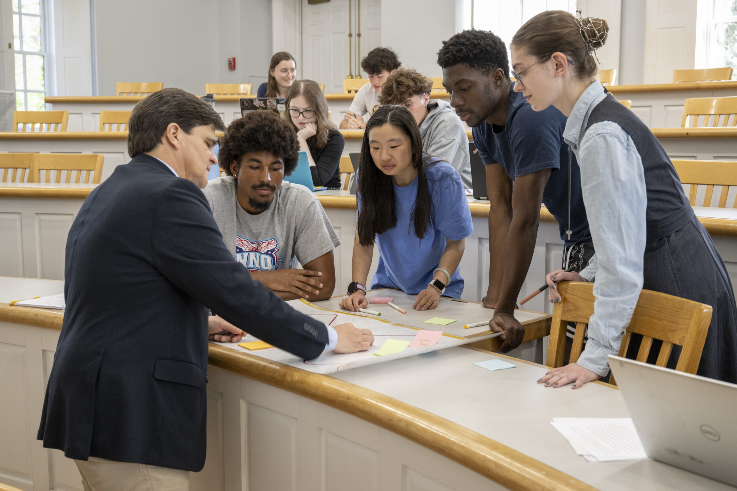 A professor and four students discuss notes and sticky notes on a table in a classroom with tiered seating and open laptops.