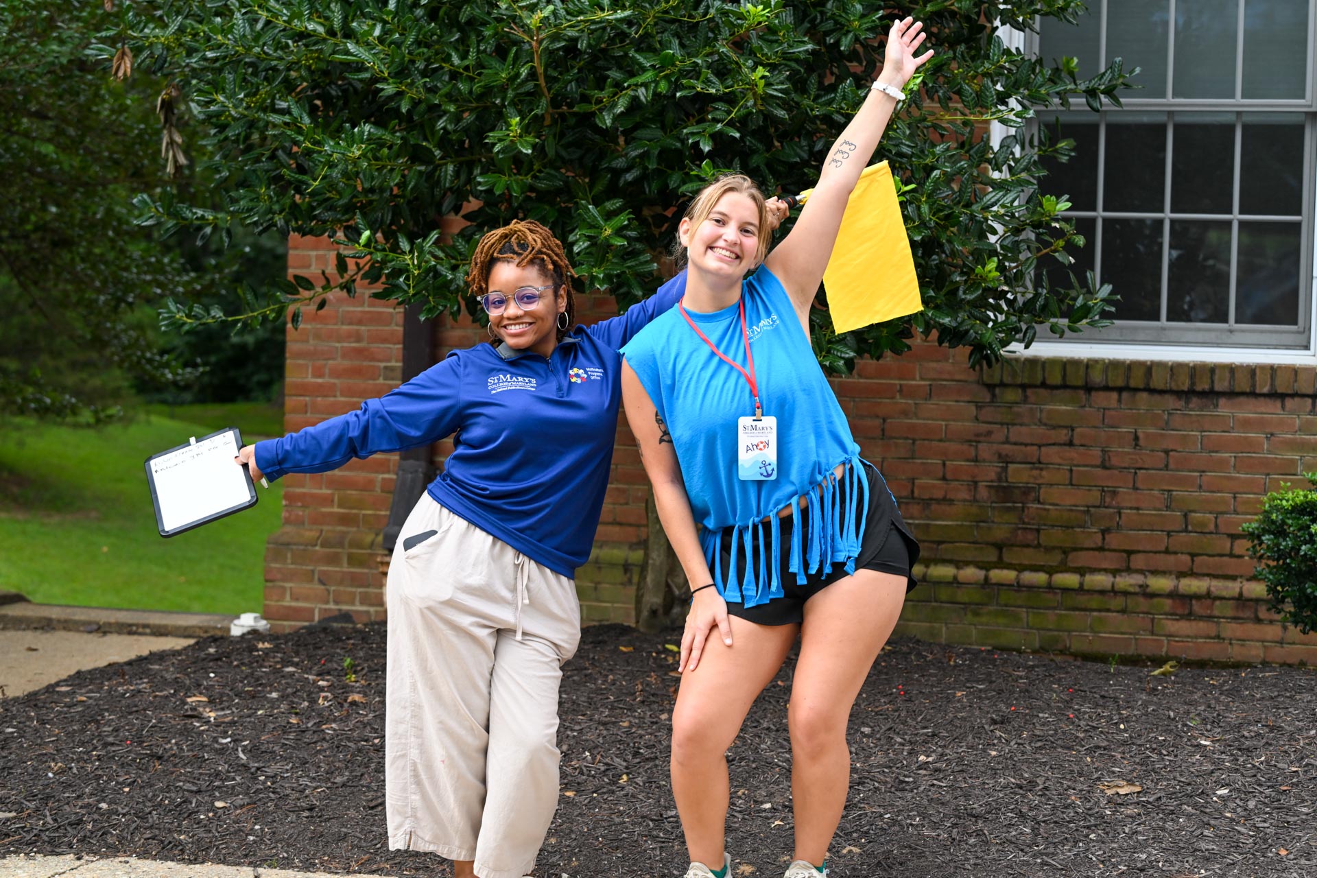Two smiling people pose outdoors in front of a brick building, one holding a clipboard and the other raising a yellow paper, both appearing cheerful and energetic.