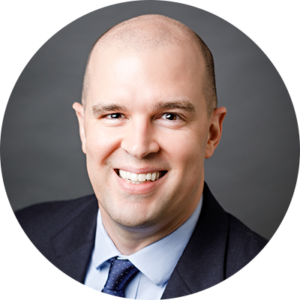 Smiling man with a shaved head wearing a dark suit, light blue shirt, and dark tie against a gray background.