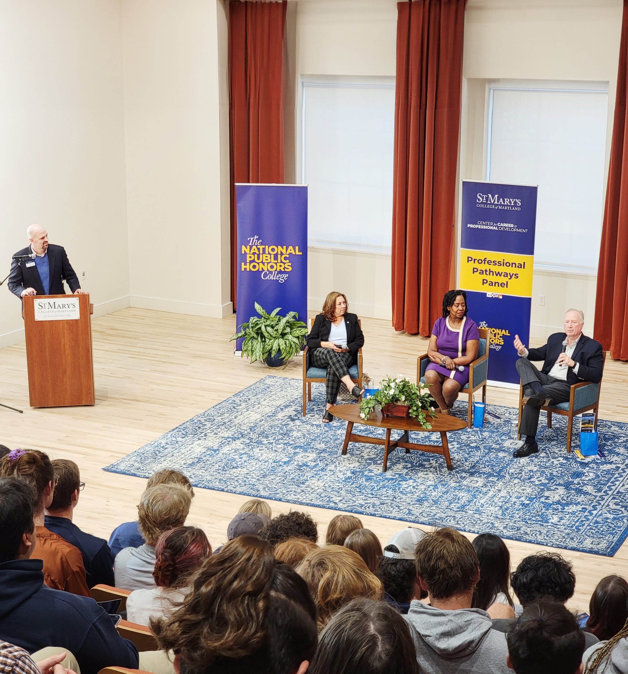 Audience watching a panel discussion with four people on stage, three seated and one at a podium. Banners display "St. Mary's College" and "Professional Pathways Panel." Attendees are seated in a room with wooden floors and large windows.