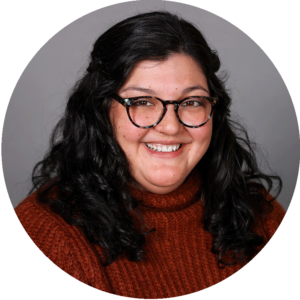 Smiling person with long curly black hair and glasses, wearing a brown sweater, against a gray background.