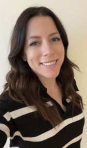 Kristen headshot A person with long, wavy dark hair smiles at the camera. They are wearing a black sweater with white stripes. The background is plain and light-colored.