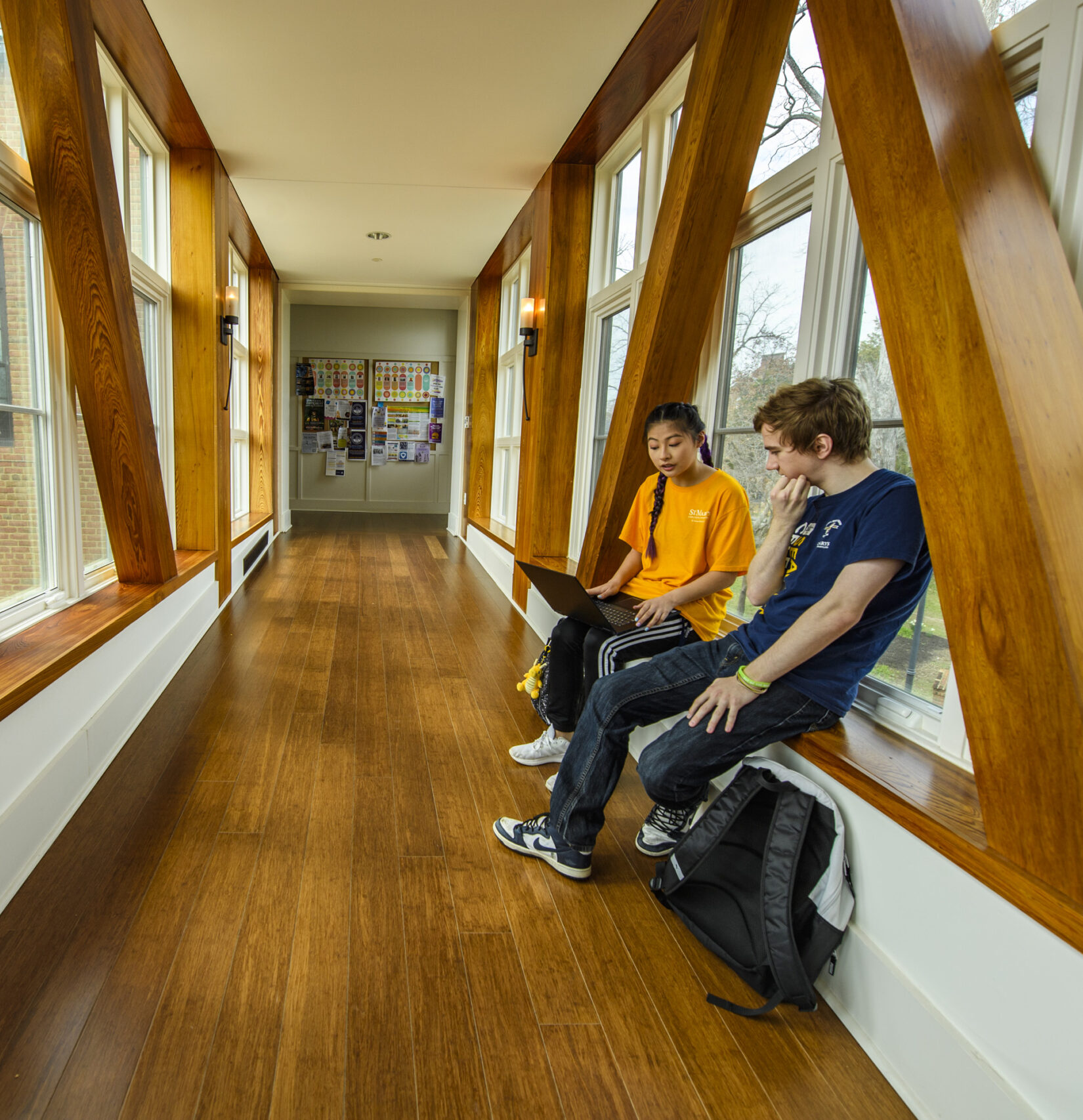 Two students sitting in a sunlit hallway with large wooden beams, chatting and looking at a laptop. A bulletin board with colorful papers is visible in the background. Backpacks are placed on the floor beside them.