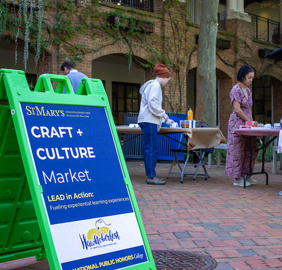 A group of people interact at tables displaying various items at an outdoor Craft and Culture Market. A green sign in the foreground reads "St. Mary's Craft + Culture Market" promoting experiential learning.