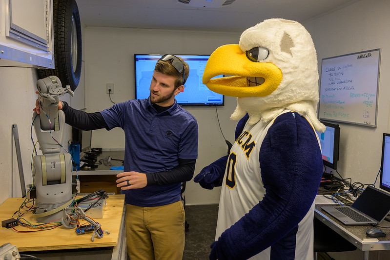 A person in a blue shirt and cap examines a robotic arm on a wooden table. Beside them, an eagle mascot in a sports jersey watches. Two computer screens and a whiteboard with notes fill the background.