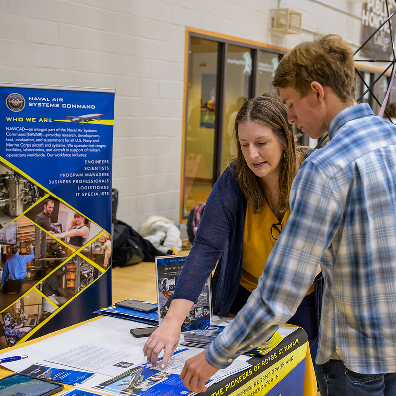 Photo by Chuck Steenburgh A woman explains information to a young man at a Navy recruitment booth. The booth has banners and brochures about Naval Air Systems Command. They are indoors, surrounded by tables and other booths.
