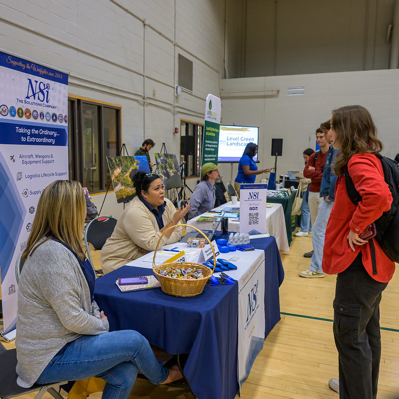 Photo by Chuck Steenburgh A career fair scene with multiple booths in a sports hall. People are conversing at tables displaying marketing materials and banners. One table has a sign "MSB The Department," with brochures and a bowl of candy. Attendees are casually dressed.