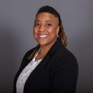 Smiling person with short dreadlocks and one side shaved, wearing a black blazer over a white polka dot blouse, stands against a gray background.