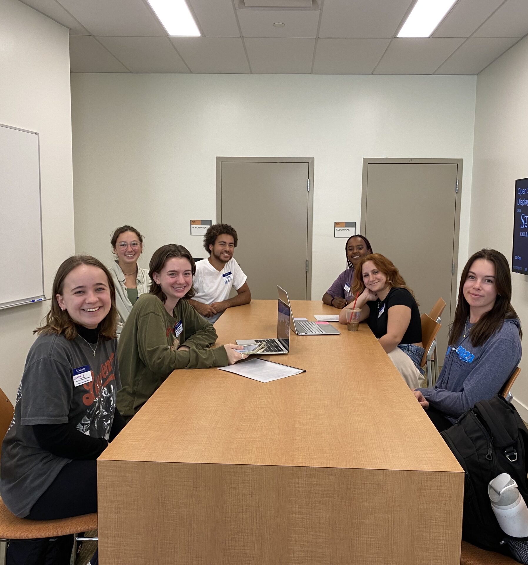 Seven people sit around a rectangular wooden table in a brightly lit room with a whiteboard and a screen displaying "St. Mary's" on the wall. They are smiling and some have laptops open.