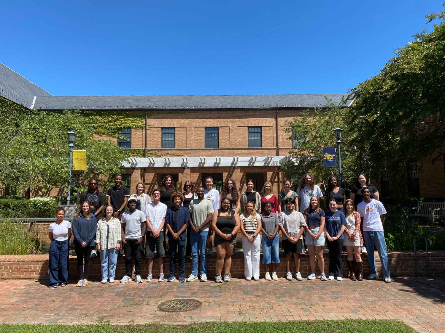 A group of about 30 people stands in two rows outside a brick building on a sunny day, posing for a group photo.