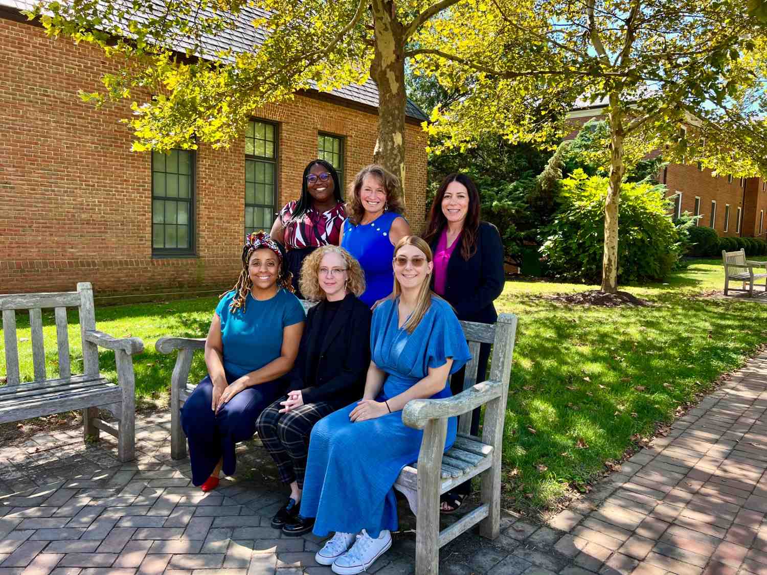 Six women pose for a group photo on and around a wooden bench outside, with trees and brick buildings in the background on a sunny day.