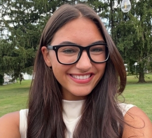 A woman with long brown hair and black glasses smiles outdoors in front of trees and grass.