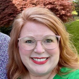 A woman with red hair, glasses, and a green top is smiling outdoors with green grass and red-leaved bushes in the background.