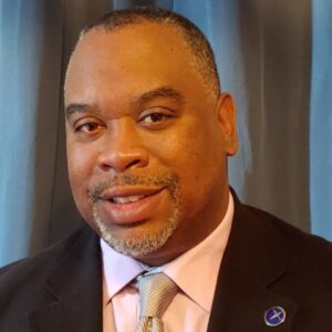 A man in a suit and tie poses in front of a blue curtain, wearing a light pink shirt and a pin on his lapel.