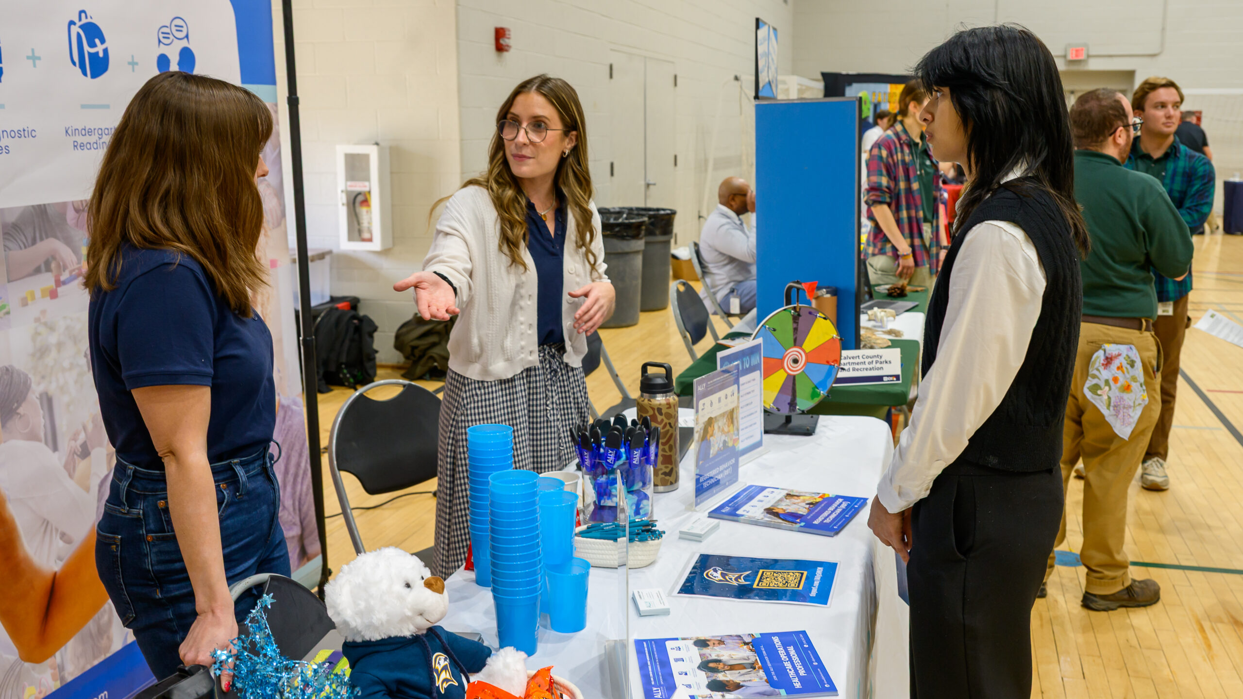 A woman at a career fair booth gestures while speaking to a visitor across a table with brochures, pens, and stacks of blue plastic cups.
