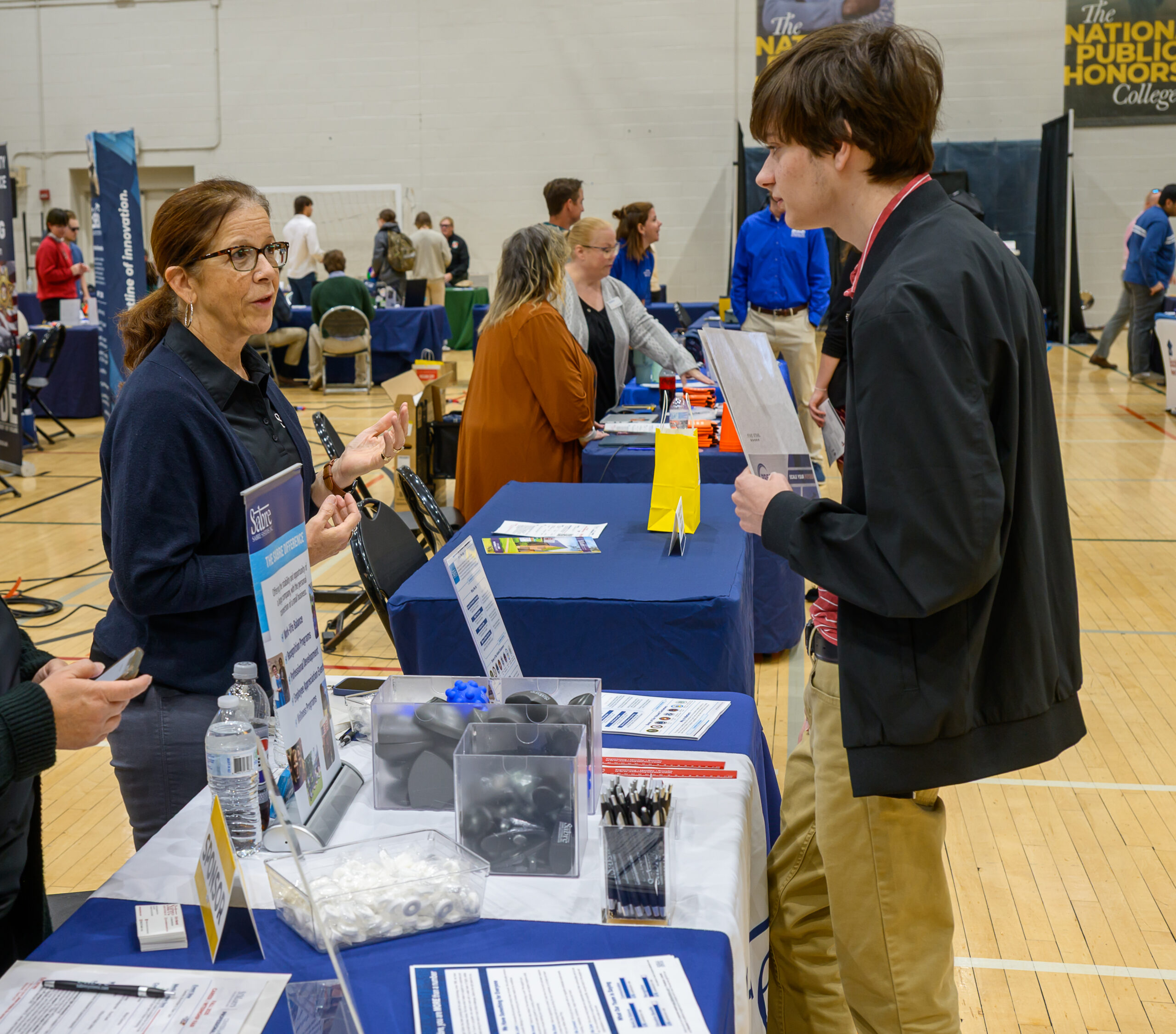 A woman speaking to a young man at a college fair booth.