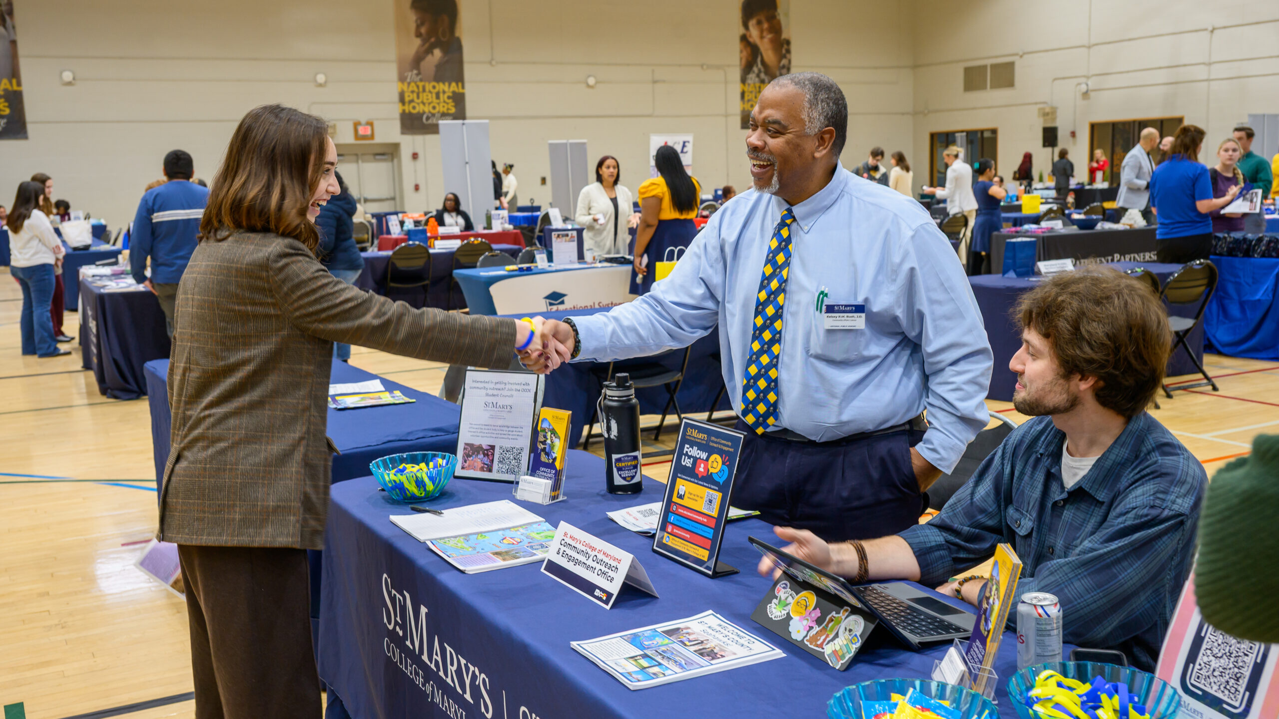 A woman in a blazer shakes hands with a man wearing a tie at a St. Mary’s College information table in a gymnasium.
