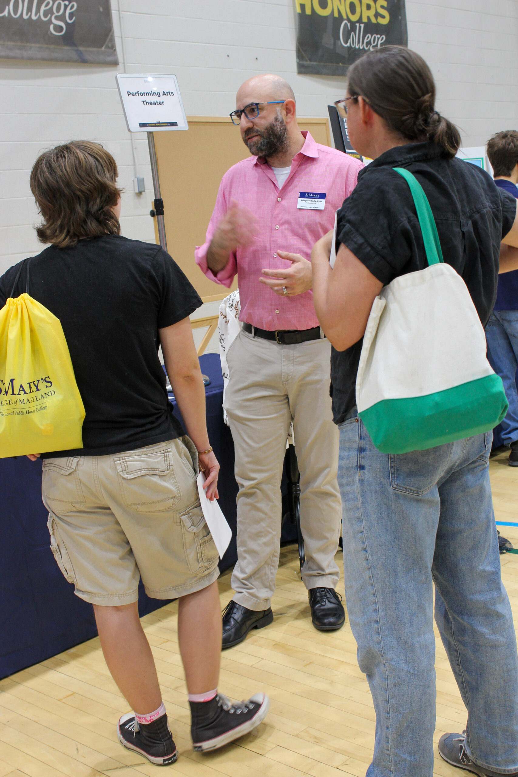 A bearded man in a pink button-down shirt talks to two students at a college fair booth.