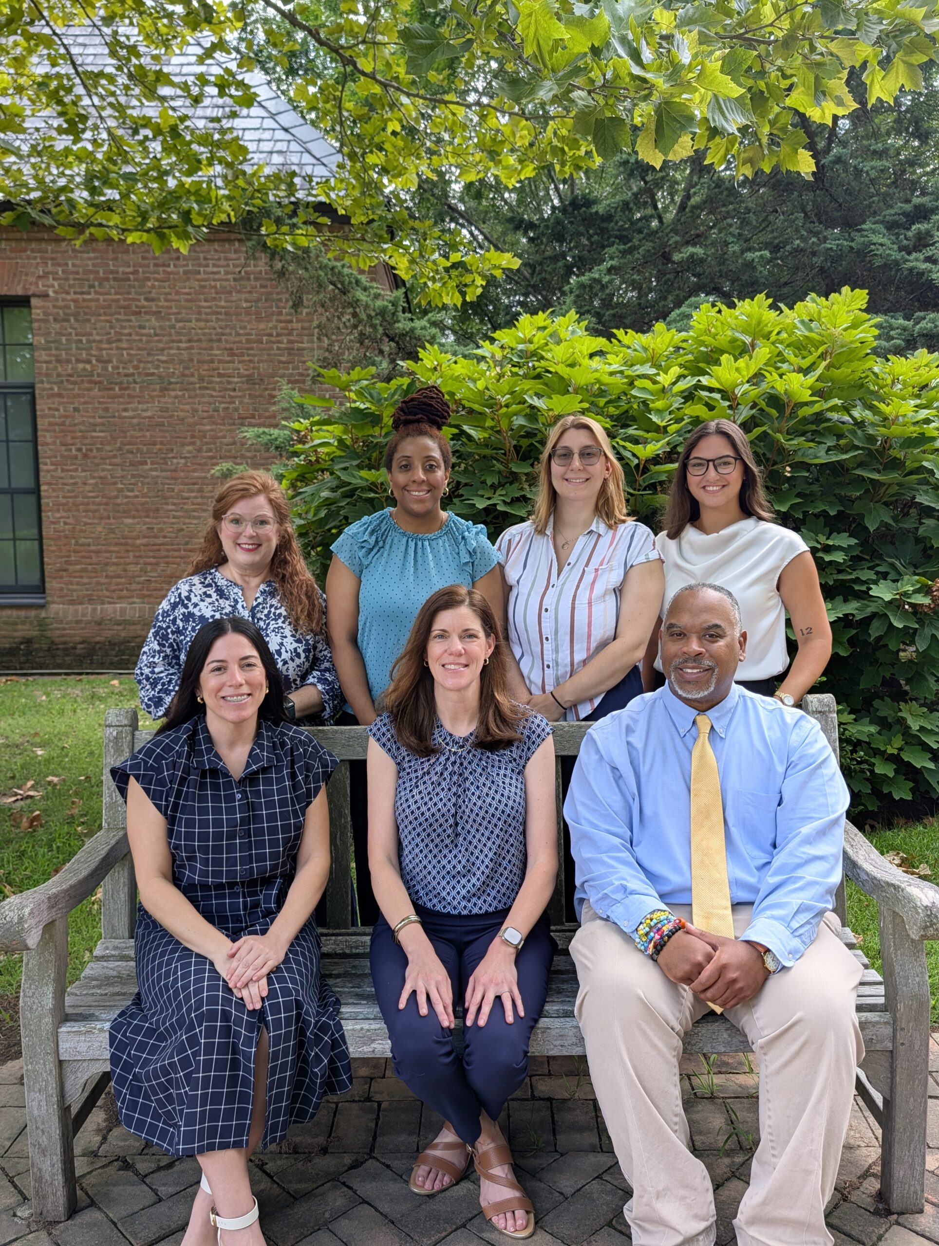 A group of seven people posing for a photo on a wooden bench outdoors.