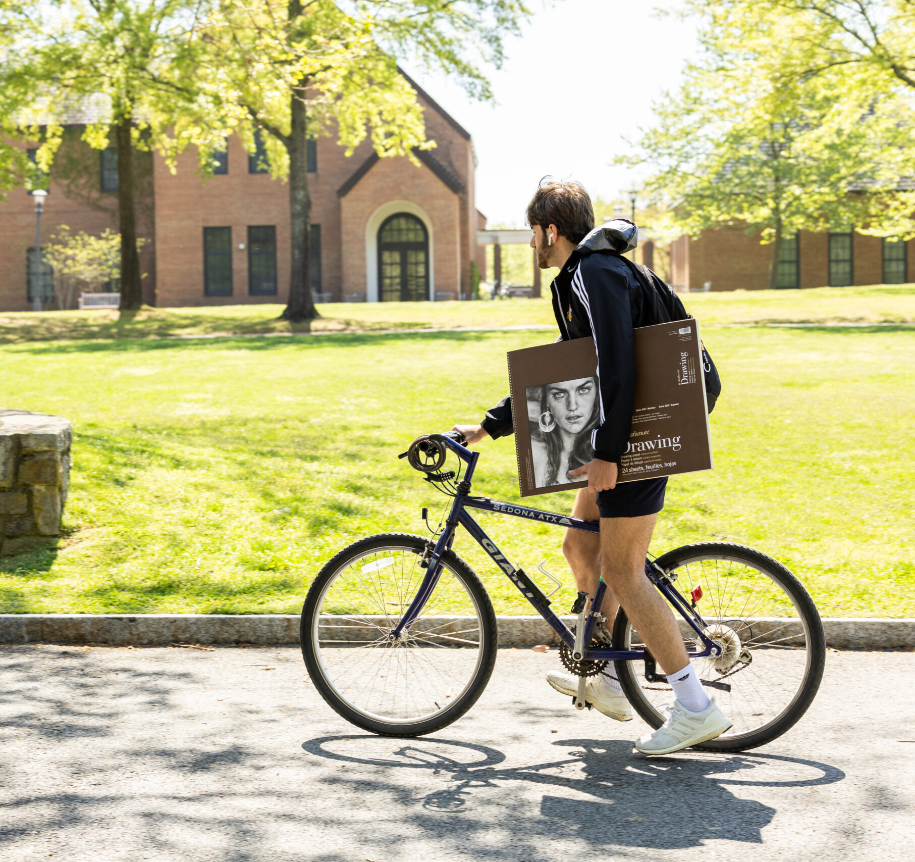 A person wearing a backpack rides a bicycle while carrying a large drawing pad with a sketch of a face on a sunny campus pathway.