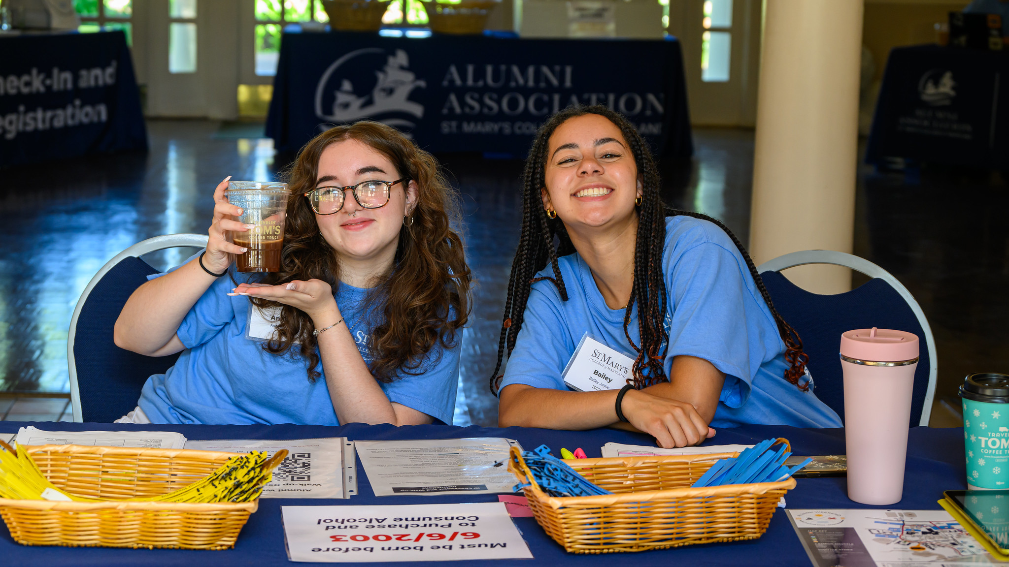 Two smiling people sit at a registration table for an alumni event, wearing blue event T-shirts. One holds up a cup, and the table is covered with papers, pens, and two baskets. A banner in the background reads "Alumni Association.