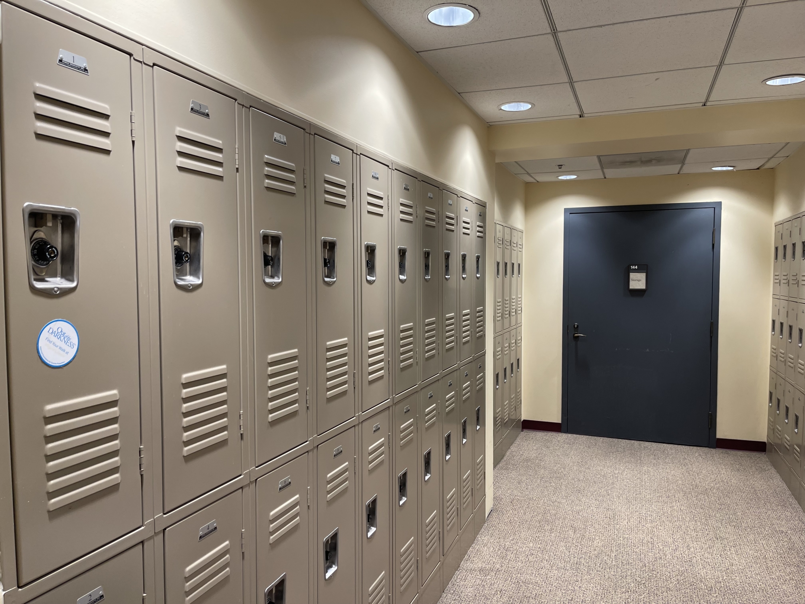 74490514813__5F3298DE-F3F9-44D7-A536-E626E2F3DA31 A hallway with beige lockers lining both sides. The ceiling has recessed lighting, and there is a dark gray door at the end of the corridor. The floor is covered with beige carpet.