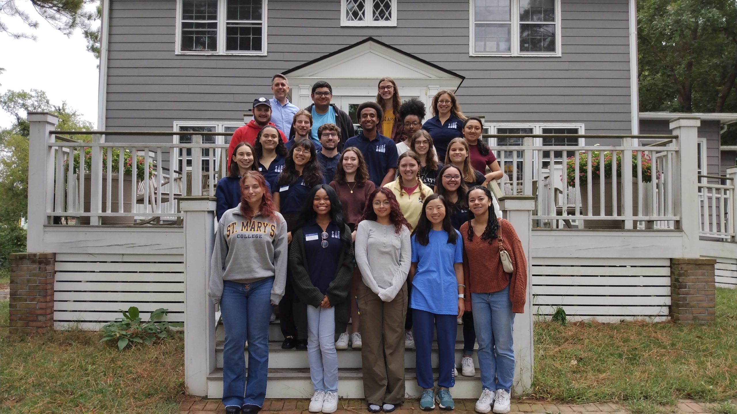 A diverse group of people stands on a wooden stairway in front of a gray house with white trim. They are smiling and dressed casually. The group includes men and women of various ages, and the background shows trees and grass.