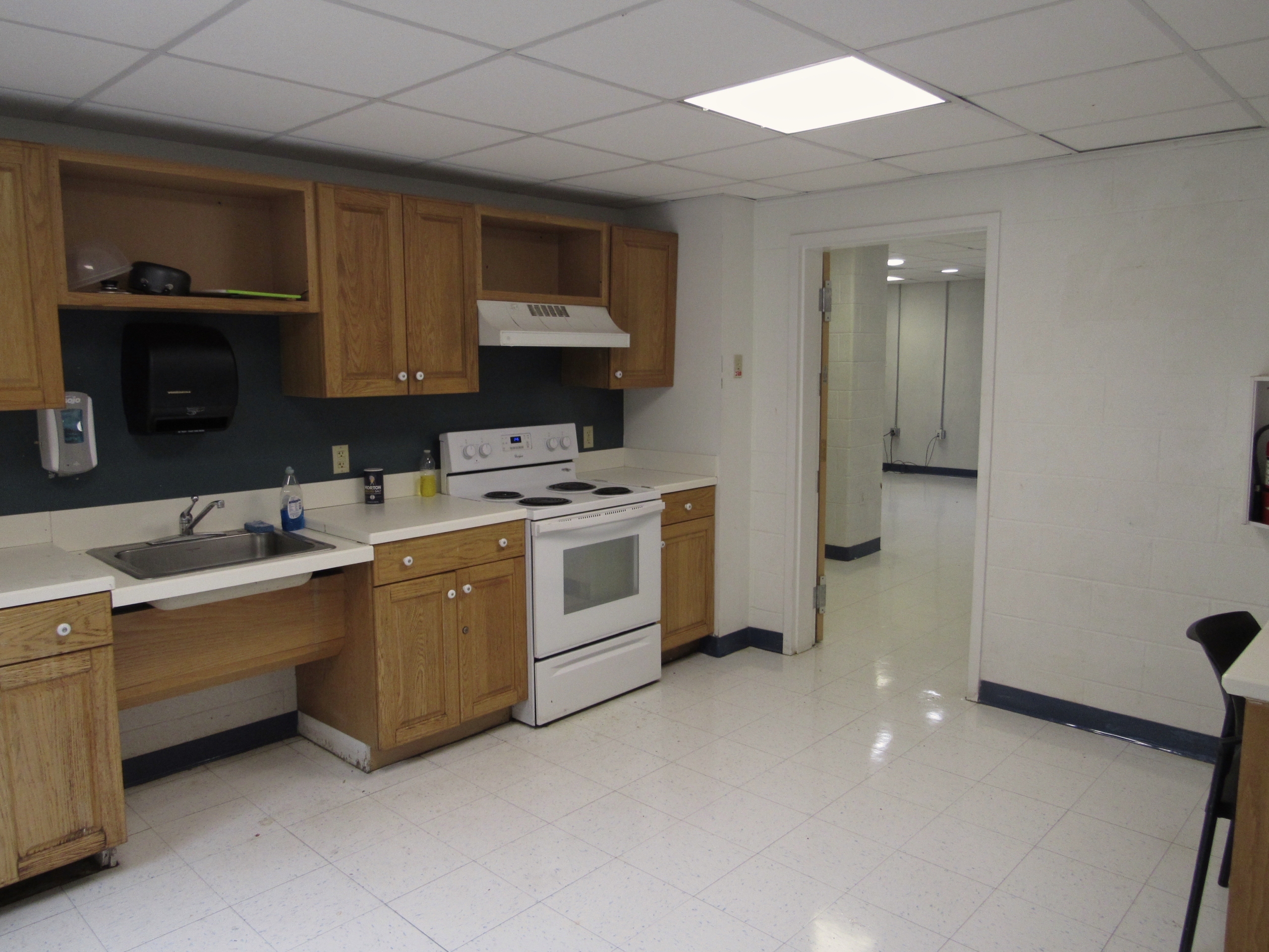 IMG_0013 A small kitchen with wooden cabinets, a white stove, and a sink. The countertops are white, and there's a paper towel dispenser on the wall. The floor is tiled, and a hallway leads to another room. The lighting is provided by ceiling panels.