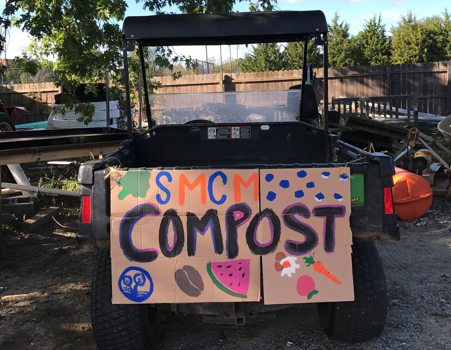 A utility vehicle with a colorful 'SMCM COMPOST' sign featuring drawings of a globe, coffee beans, and produce stands outdoors by trees and a fence.