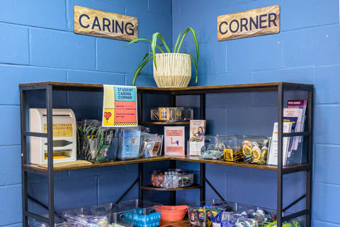 A metal shelf in a blue-walled corner labeled "Caring Corner." It holds supplies like snacks, hygiene products, and stationery. A plant sits on top, and a sign reads "Student Corner." Brochures and leaflets are also displayed.