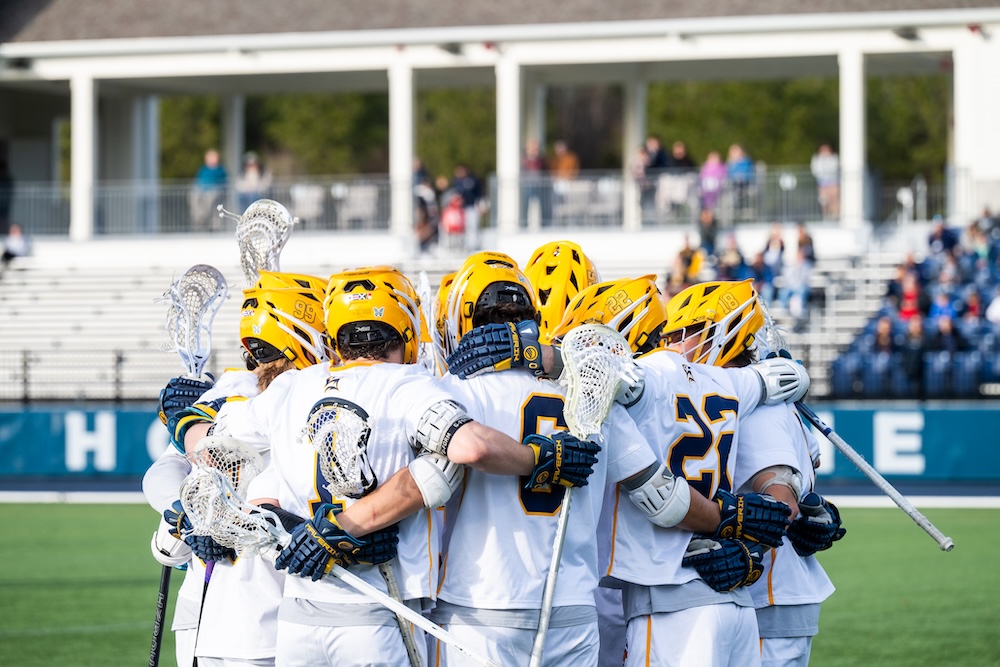 A lacrosse team in yellow helmets and white uniforms huddles together on a field before a game, with spectators seated in the background.