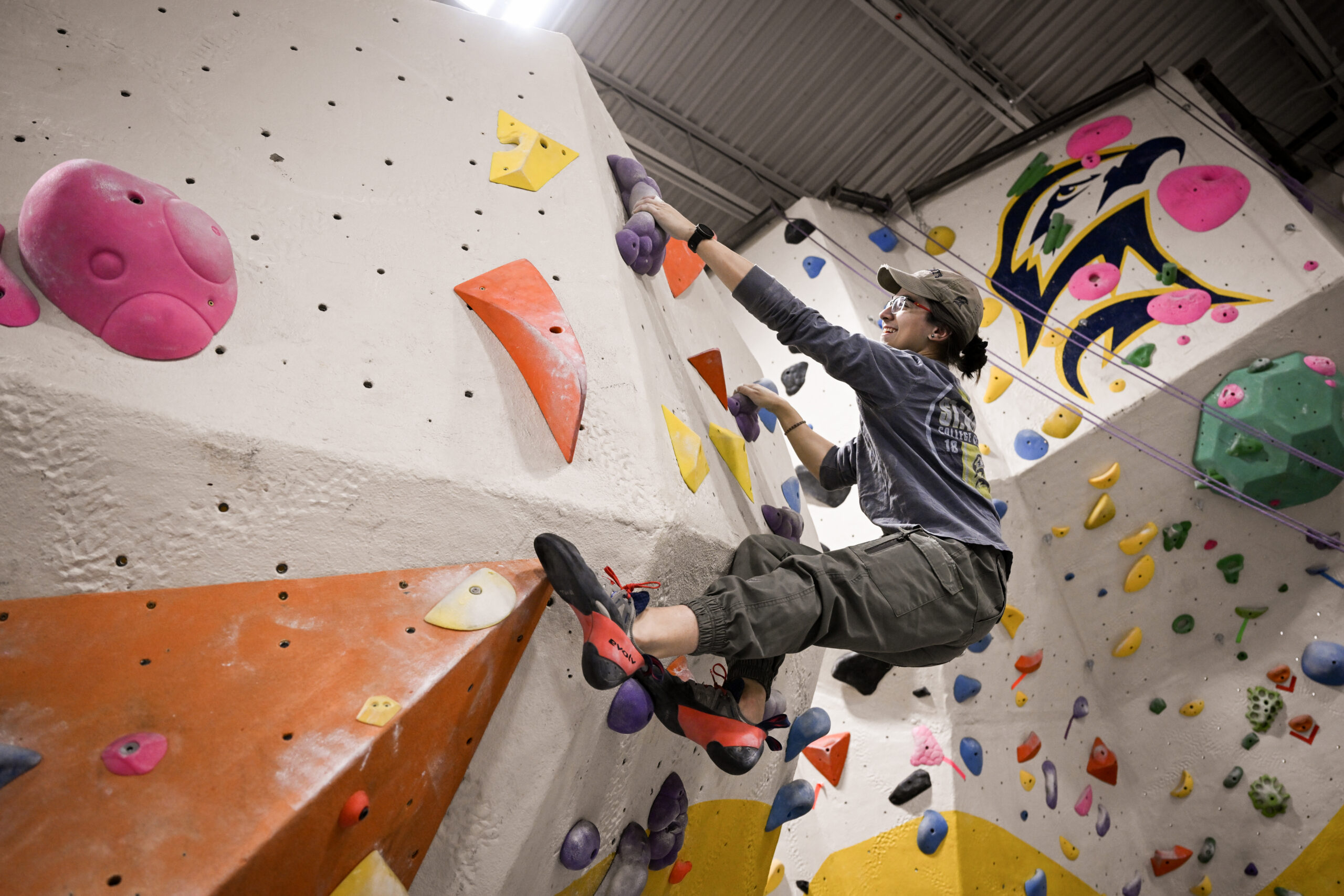 A person wearing a hat and climbing shoes ascends an indoor bouldering wall with colorful holds and geometric features.