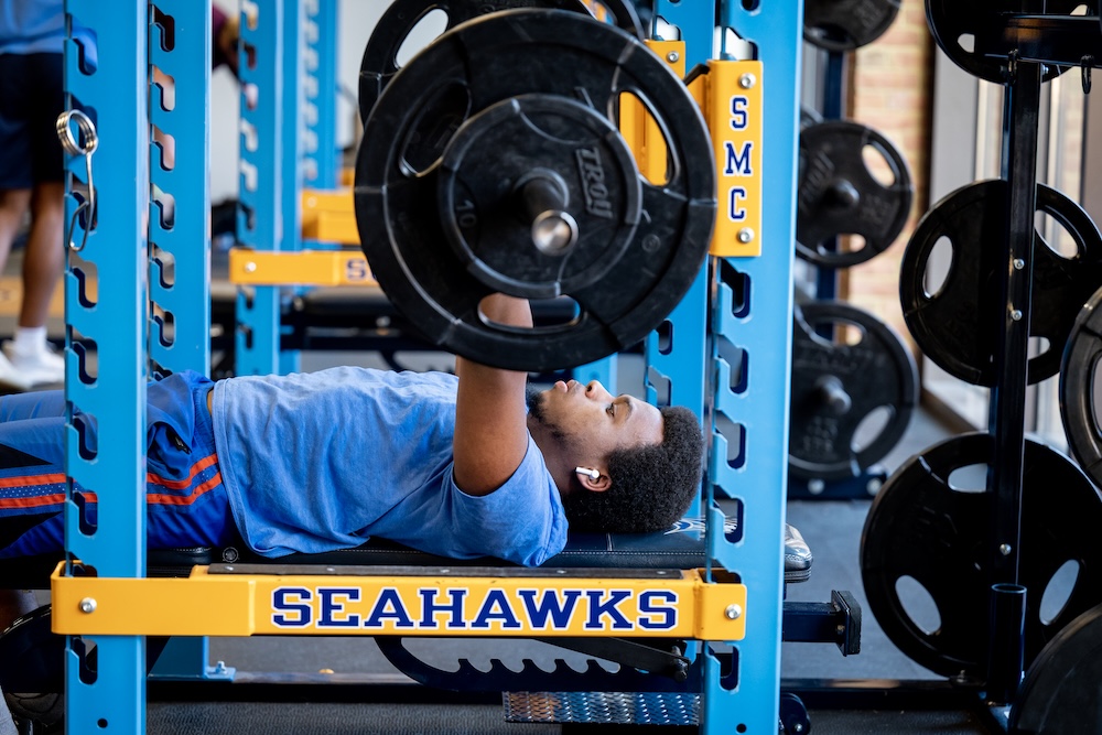 A person in a blue shirt is bench pressing weights on a rack labeled "SEAHAWKS" in a gym. Weight plates are visible in the background.