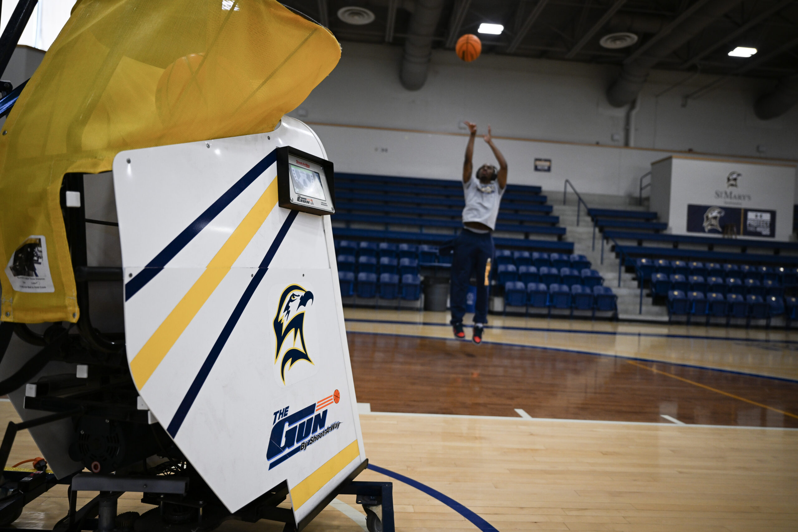 A basketball player practices shooting in a gym while a basketball shooting machine collects and returns balls on the court.