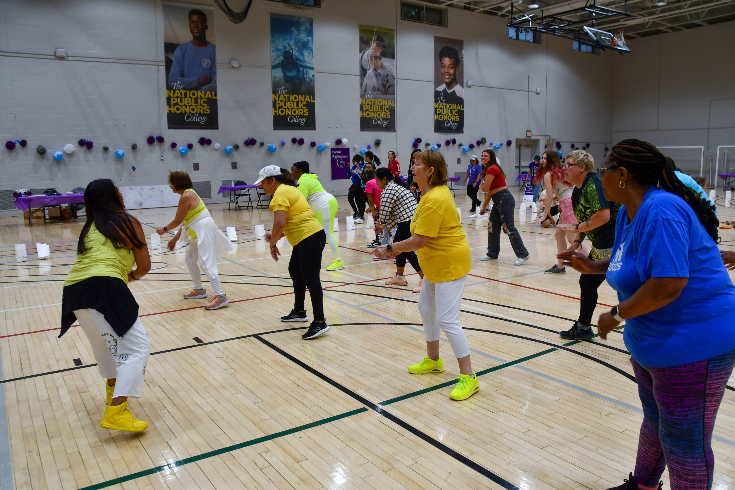 A group of adults exercise together in a gymnasium, following an instructor. Some participants wear yellow shirts, and the gym is decorated with balloons and banners.
